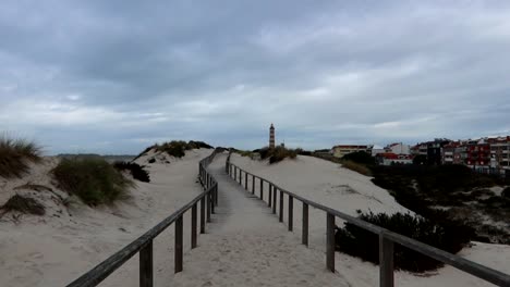 Sandy-wooden-path-leads-to-Costa-Nova-Beach-with-Barra-Lighthouse,-cloudy-sky-over-coastal-dune