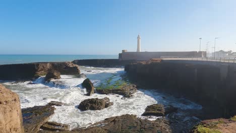 Waves-crash-into-rocks,-splashing-on-cliff,-Rabat-lighthouse-on-shore-of-the-Atlantic-Ocean