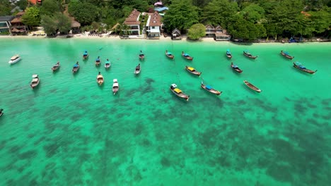 Thailand-Long-Tail-Boats-anchored-by-beach-in-beautiful-blue-tropical-water--aerial-fly-in