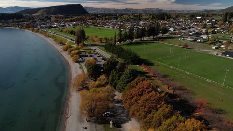 Drone-fly-over-lake-Wanaka-shore-and-recreation-area-to-the-town,-during-Autumn-season