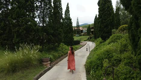 Lady-in-Red-Walking-on-a-Path-in-an-Italian-Styled-Botanical-Garden-Resort-with-Surrounding-Scenery-in-Ratchaburi,-Thailand
