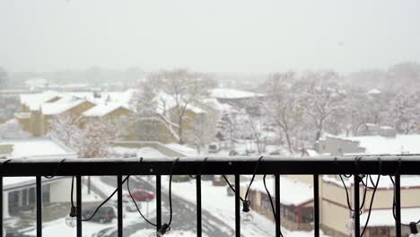 Medium-shot-of-snowflakes-falling-during-a-old-winter-snowstorm-with-an-apartment-balcony-railing-and-other-buildings,-trees,-and-a-parking-lot-out-of-focus-in-the-background-in-Midvale-Utah