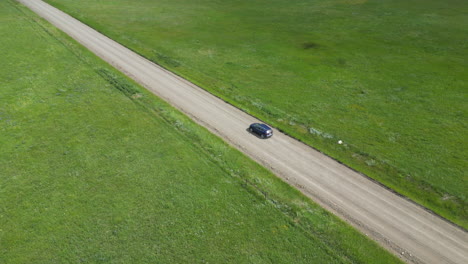 Blue-Car-Speeds-Down-Dirt-Road-with-Green-Grass-Fields-on-Either-Side-of-the-Trail
