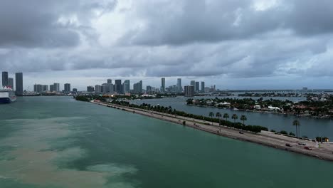 Una-Transitada-Carretera-Rodeada-De-Agua-En-El-Popular-Puerto-De-Cruceros-De-Miami,-Florida,-Con-Suciedad-En-El-Agua-Del-Barco-En-Movimiento-Y-Un-Crucero-Y-Rascacielos-Al-Fondo-En-Un-Cálido-Día-Nublado