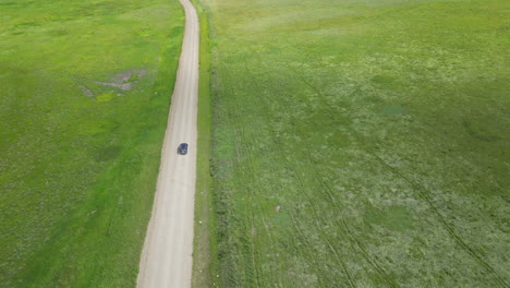Aerial-View-of-Car-Traveling-Down-Dirt-Road-with-Stunning-Green-Grass-Surrounding-It