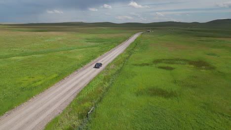 Blue-SUV-Car-Driving-Down-Dirt-Road-in-the-Middle-of-Green-Grassy-Pasture