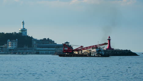 Grab-Dredger-Crane-Ship-Cruising-In-The-Ocean-Passing-By-On-Red-Lighthouse-with-Sokcho-Lighthouse-In-The-Distance