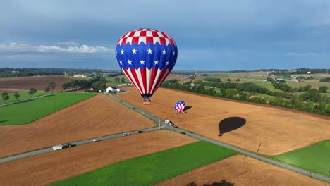 Luftaufnahme-Eines-Fliegenden-Heißluftballons-Mit-Amerikanischer-Flagge-Und-Sternen-über-Dem-Ländlichen-Amerikanischen-Gebiet-Und-Fahrenden-Autos-Auf-Der-Straße