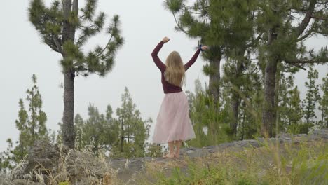 Joyful-blonde-haired-woman-in-long-pink-skirt-black-top-swings-arms-happy-and-barefoot,-rear-view