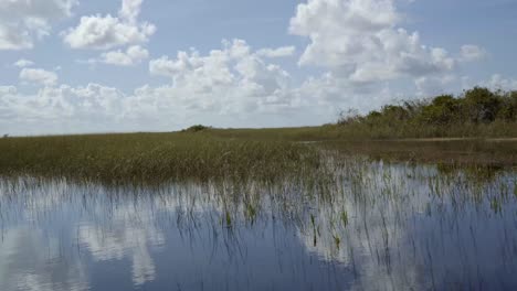 Fast-trucking-left-shot-of-the-stunning-Florida-everglades-near-Miami-riding-on-a-airboat-with-the-calm-swamp-water-reflecting-the-sky-and-creating-a-mirage-surrounded-by-tall-grass-on-a-sunny-day