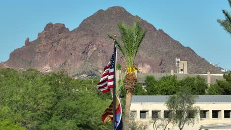 Banderas-De-Estados-Unidos-Y-Del-Estado-De-Arizona-Ondeando-Frente-A-Una-Palmera-Y-Un-Fondo-De-Montaña