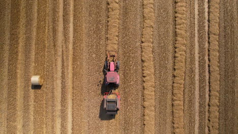Top-down-aerial---descending-towards-a-tractor-with-a-baling-machine-standing-in-a-field-next-to-a-huge-bale-of-straw