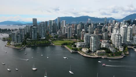 Aerial-view-showing-many-boats-on-Falls-Creek-River-and-gigantic-Skyscraper-Tower-of-Vancouver-City-in-background---Mountain-range-silhouette-in-backdrop---Rising-drone-flight