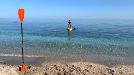 Girl-slowly-puddle-surfing-in-the-sea-in-Nerja-Malaga,-stand-up-puddle-boarding-on-a-sunny-day-in-Spain,-fun-vacation-activity,-adventurous-water-sport,-4K-static-shot