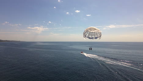 Aerial-view-of-Parasailing-in-Marbella-Malaga,-fun-water-activity-on-a-sunny-day,-fast-boat-pulling-Mistral-beach-parachute-with-two-people-in-Spain,-4K-shot
