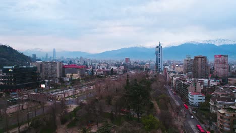 Flyover-the-Forestal-park-on-a-cloudy-morning-with-the-snowy-Andes-mountain-range-in-Santiago-Chile,-telephone-tower-and-light-pollution-in-the-background