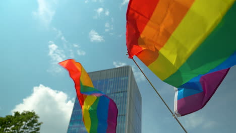 Flags-in-LGBT-colors-flutter-in-the-wind,-mounted-on-wooden-and-plastic-poles---in-the-background,-a-modern-glass-building