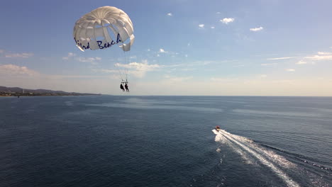 Aerial-view-of-Parasailing-in-Marbella-Malaga,-fun-water-activity-on-a-sunny-day,-fast-boat-pulling-Mistral-beach-parachute-with-two-people,-blue-sky-in-Spain,-4K-shot