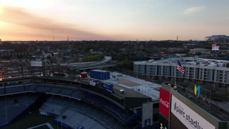 Drone-circles-around-empty-Center-Parc-Stadium-with-the-American-flag-waving-at-sunset,-Downtown-Atlanta-street,-and-skylines-in-the-background