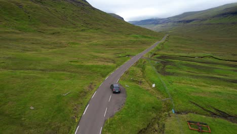 Aerial-following-shot-of-a-stopped-lone-car-starting-driving-between-green-Faroese-mountains