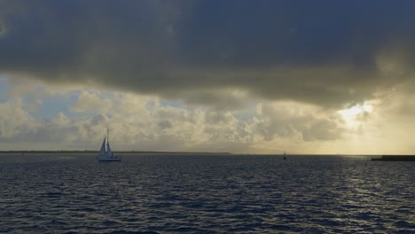 Desde-La-Perspectiva-De-Un-Barco,-Una-Toma-Fascinante-De-Enormes-Nubes-Al-Atardecer,-Bañadas-Por-Rayos-Divinos,-Con-Un-Velero-Navegando-Tranquilamente-Cerca