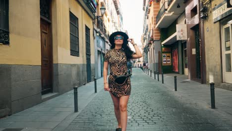 Girl-walking-down-the-streets-of-Spain,-old-style-buildings,-chetah-dress-on,-with-black-hat