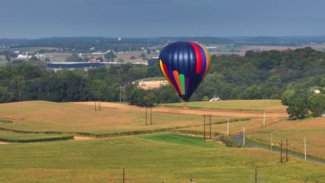 Bunter-Heißluftballon,-Der-Langsam-über-Den-Rand-Der-Amerikanischen-Stadt-In-Den-Grünen-Feldern-Fliegt,-Mit-Himmel-Im-Hintergrund,-Luftdrohnenaufnahme