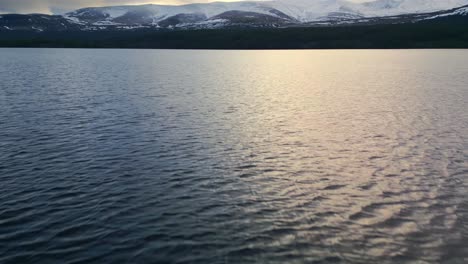 Aerial-Drone-Footage-Begins-Overlooking-a-Tranquil-Scottish-Lake,-Ascending-to-Reveal-a-Mesmerizing-Panorama-of-Majestic-Snow-Clad-Mountains-and-Vast-Highland-Terrain