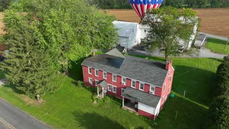Ascending-drone-shot-of-house-in-american-countryside-and-hot-air-balloon-landing-on-agricultural-field-in-background---rising-aerial-shot