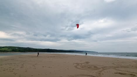 Active-kitesurfing-couple-preparing-kiteboarding-water-sports-equipment-on-windy-sandy-Wales-beach,-Wide-shot