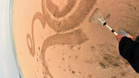 Vertical-POV-male-using-rake-to-create-swirling-zen-sand-design-patterns-on-golden-Anglesey-beach