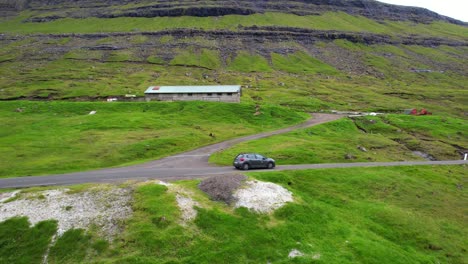 Drone-tracking-a-car-driving-to-Saksun-in-a-road-surrounded-by-mounts-and-waterfalls-in-Faroe-islands