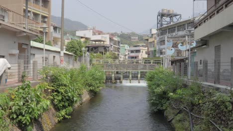 Ein-Fluss,-Der-Durch-Einen-Kanal-Mit-Dreistöckigen-Gebäuden-Auf-Beiden-Seiten-Und-Einer-Kleinen-Dammbrücke-Im-Hintergrund-In-Einem-Kleinen-Dorf-Im-Süden-Taiwans-Fließt-–-Totale