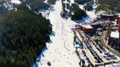 Bird's-Eye-View-of-a-Ski-Trail-with-People-Riding-Chairlifts-down-Slope-next-to-Parking-Lot-and-Pine-Tree-Forest-in-Snow