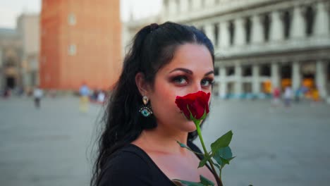 Girl-holding-red-rose,-close-up-shot,-panning-around-girl,-black-hair,-black-dress,-in-city,-buildings-in-background
