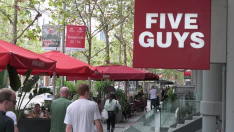 Pedestrians-walk-past-the-American-fast-food-casual-restaurant-chain-Five-Guys-and-its-logo-sign-in-Madrid,-Spain