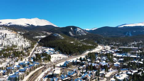 Luftdrohnenaufnahme-Von-Schneebedeckten-Häusern-Und-Bergen-In-Der-Wunderschönen-Bergstadt-Breckenridge,-Colorado,-Mit-Menschen,-Die-Auf-Der-Straße-Durch-Kiefernwälder-Fahren
