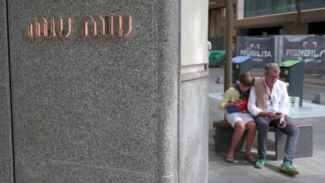 A-father-and-a-son-sit-on-a-bench-outside-the-Italian-high-fashion-women's-clothing-and-accessory-brand-Miu-Miu-store