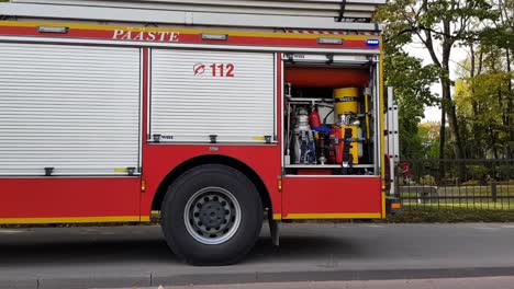 Rotes-Scania-Feuerwehrauto-Mit-Eingeschalteten-Lichtern-Neben-Der-Straße.-Autos-Fahren-Vorbei