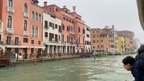 Los-Turistas-Disfrutan-De-La-Impresionante-Vista-De-Los-Barcos-Flotando-En-El-Agua,-Mientras-Que-Los-Encantadores-Y-Vibrantes-Edificios-Realzan-Aún-Más-El-Encanto-De-La-Calle-Del-Agua-De-Venecia.
