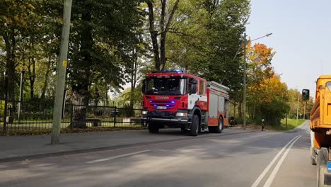 Rotes-Scania-Feuerwehrauto-Mit-Eingeschalteten-Lichtern-Neben-Der-Straße.-Autos-Fahren-Vorbei