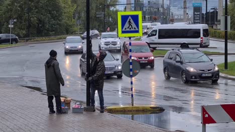 Workers-work-in-rain-next-to-busy-road-at-crosswalk,-cleaning-lamp-posts-going-around-it