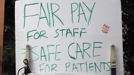 Tree-branches-and-leave-blow-in-the-window-behind-a-placard-that-reads,-“Fair-pay-for-staff-=-safe-care-for-patients”-on-a-picket-line-by-striking-consultants-and-junior-doctors