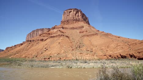 Tilt-up-handheld-wide-landscape-shot-revealing-a-stunning-orange-sandstone-rock-mountain-peak-with-the-Colorado-river-below-near-Moab,-Utah-on-a-warm-sunny-spring-morning