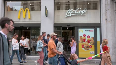 People-walk-past-the-American-multinational-fast-food-hamburger-restaurant-chain,-McDonald's,-and-its-logo