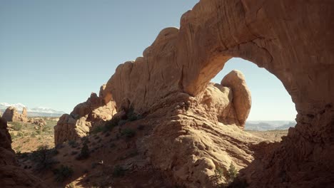 Pan-right-handheld-wide-landscape-shot-of-large-stunning-natural-orange-sandstone-arch-formations-with-sun-shining-through-them-on-a-hike-in-southern-Utah-on-a-sunny-warm-spring-morning