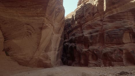 Tilt-down-handheld-shot-revealing-a-wide-opening-surrounded-by-orange-sandstone-walls-leading-to-a-narrow-dark-entrance-to-the-buckskin-gulch-slot-canyon-in-southern-Utah-near-Arizona-in-spring