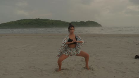 A-woman-practices-a-harmonious-blend-of-exercise-and-yoga-at-the-beach,-specifically-engaging-in-deep-squats