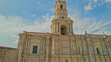 Upward-drone-pan-of-Arequipa-Cathedral's-left-tower-on-a-cloudy-day-with-the-sun-illuminating-the-tower,-a-mix-of-sun-and-clouds