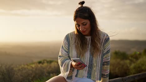 Schöne-Frau-Sitzt-Und-Benutzt-Ihr-Smartphone-Im-Freien-Mit-Sonne-Im-Hintergrund-Bei-Sonnenuntergang